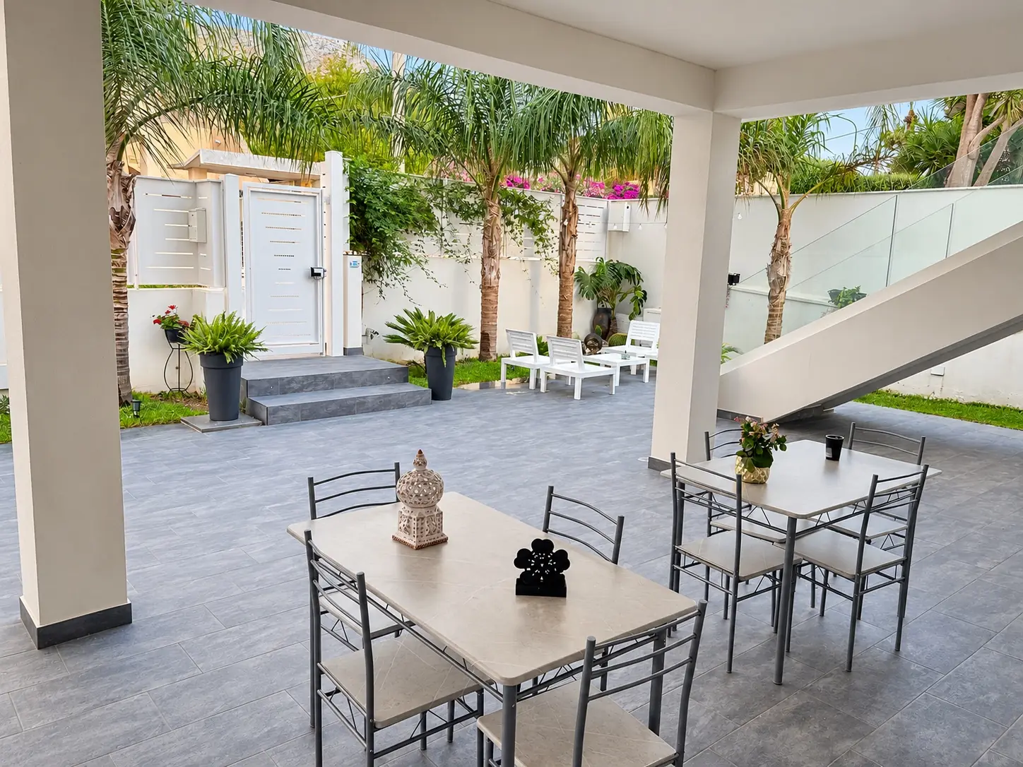 Il portico di giorno, con i due tavoli da quattro posti, le sedie e le palme del giardino sullo sfondo