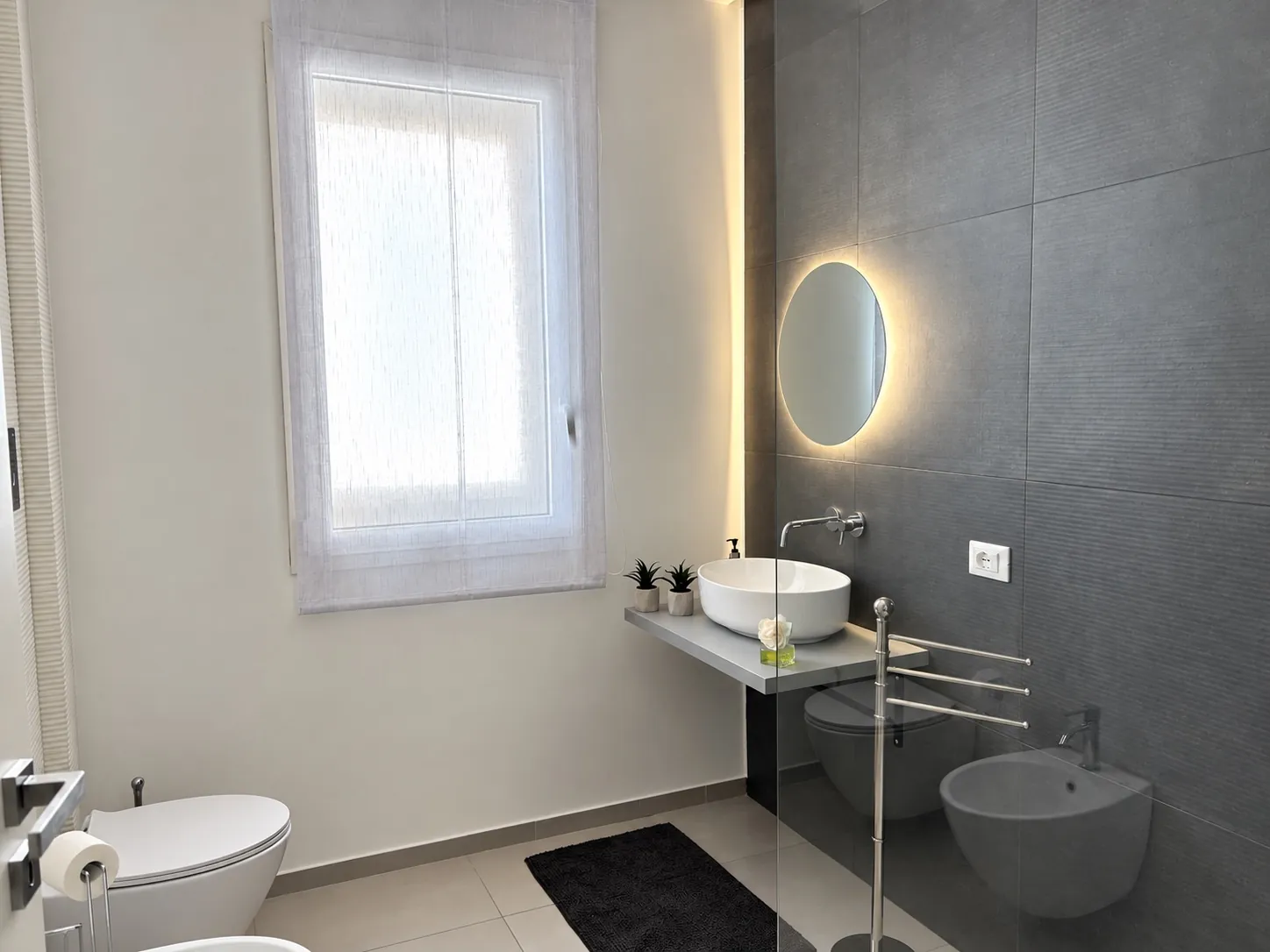 Il bagno dell'appartamento con la finestra, il bidet, il lavabo e lo specchio tondo illuminato