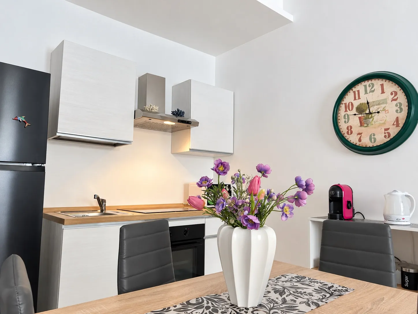 La cucina dell'appartamento vista dal tavolo da pranzo, con il vaso di fiori, il frigorifero e il bollitore