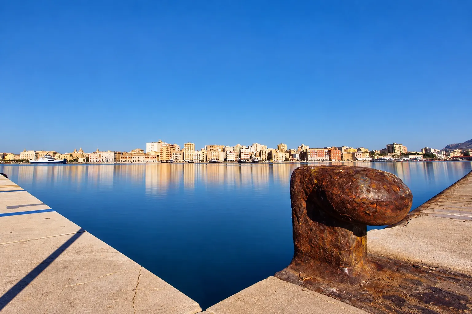 Il molo del porto di Trapani al mattino, con una bitta in primo piano e la città sul mare